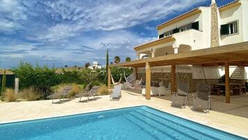 A swimming pool with lounge chairs and a pergola, surrounded by a building with a stone facade and a clear blue sky.