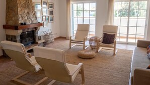 Ein Wohnzimmer mit Kamin aus Stein, zwei beigen Sesseln, einem Sofa und einem großen Fenster mit Vorhängen.