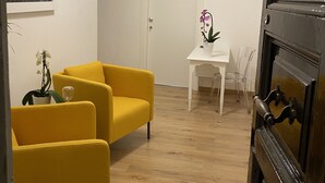 A room with a yellow sofa, a small white table with a potted orchid, a clock on the wall, and a door with a black handle. - Eden Luxury Suites (Rome)