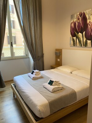 A hotel room with a bed, a painting of tulips on the wall, a window with curtains, and a nightstand with a lamp. - Eden Luxury Suites (Rome)
