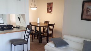 A living room with a white sofa, a dining table with chairs, a small kitchen area with a stove, and a hanging light fixture.