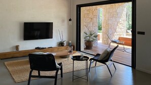 Een ruime woonkamer met een flatscreen-tv, twee stoelen, een salontafel en een groot raam dat uitkomt op een buitengebied met een stenen muur en een stoel.