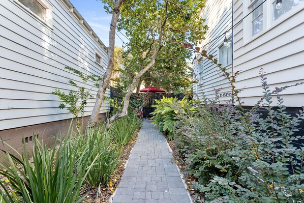 Un étroit sentier pavé de briques, bordé d’une végétation luxuriante et de bâtiments blancs.