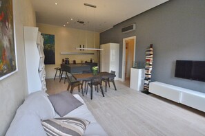 A modern living room with a sofa, dining table, and chairs. There is a TV mounted on the wall and a bookshelf with books.