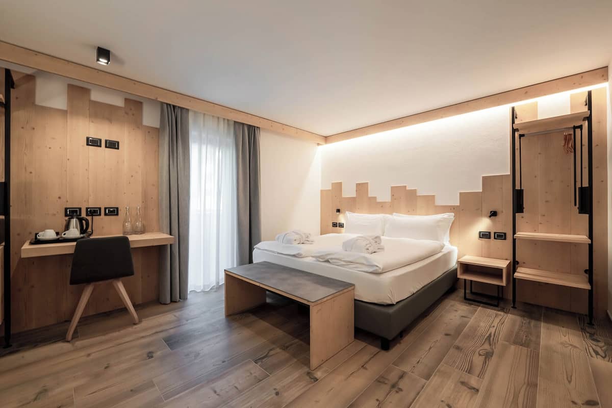 A modern hotel room with a wooden desk, a bed with white linens, a bench, and built-in shelving.