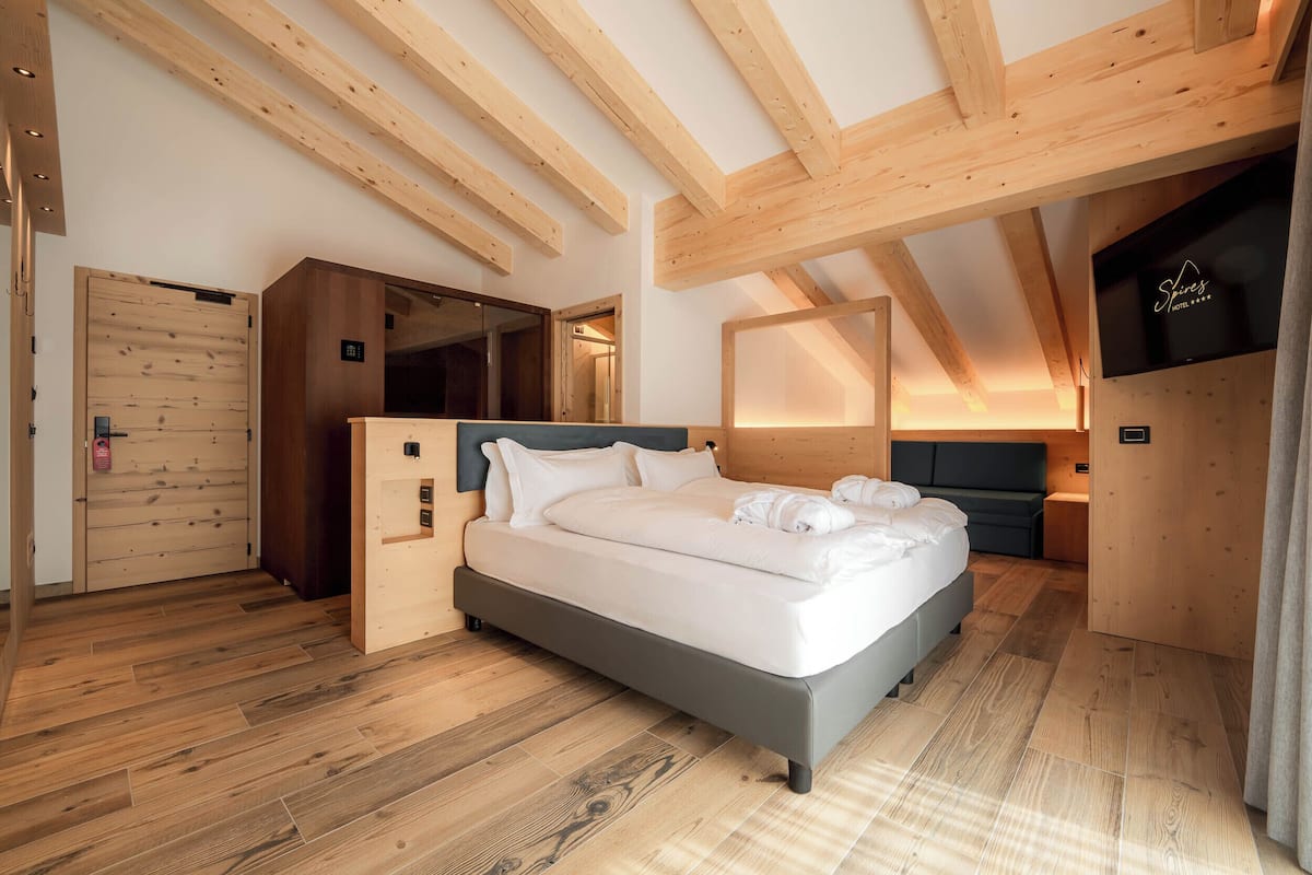 A modern hotel room with a large bed, wooden beams, and a flat-screen TV.