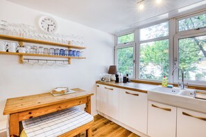 Une cuisine avec des étagères en bois, une horloge, un banc et une fenêtre donnant sur les arbres.