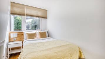 A bedroom with a bed, a nightstand, a window with blinds, and a view of greenery outside.