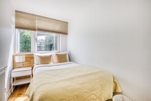 A bedroom with a bed, a nightstand, a window with blinds, and a view of greenery outside.