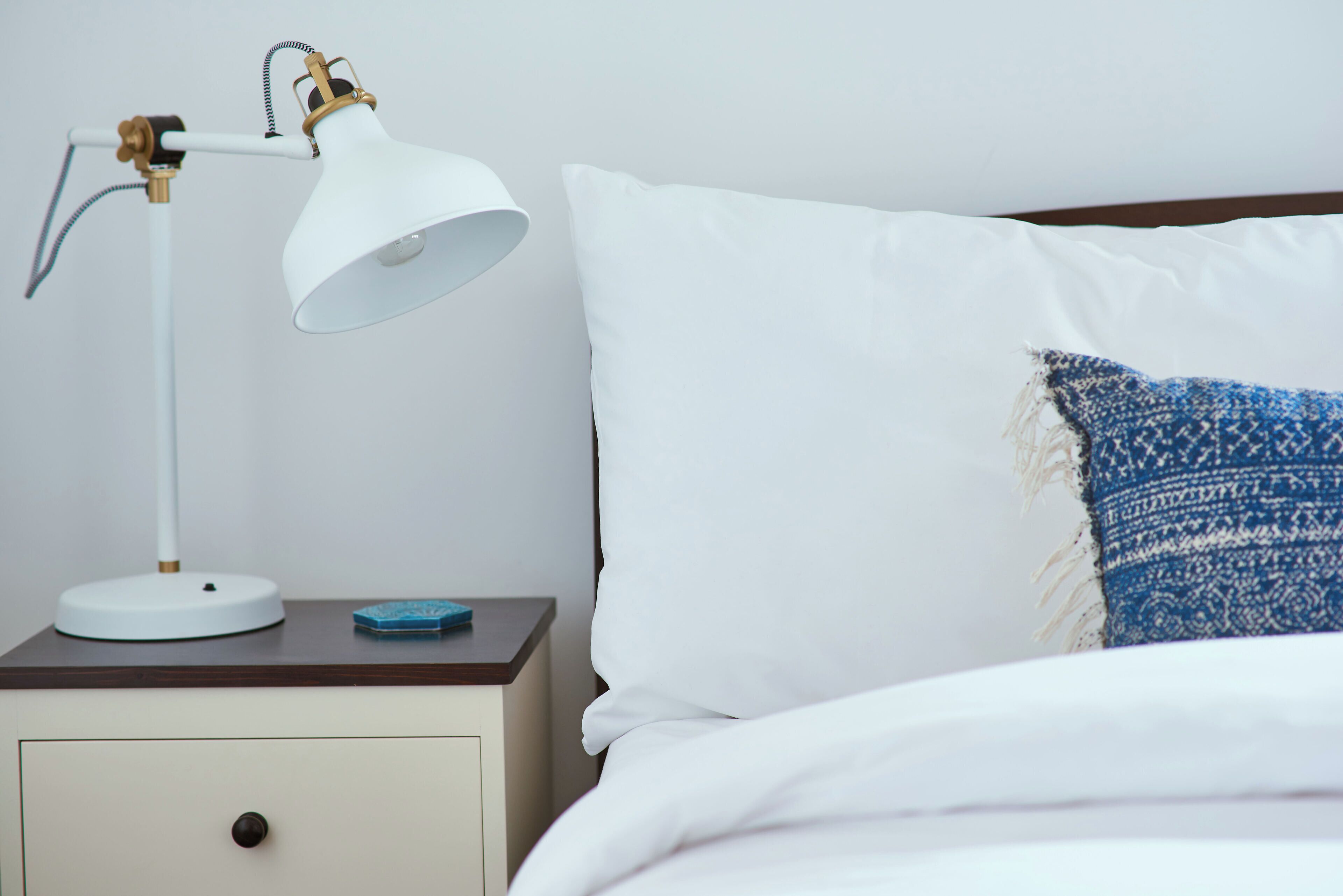 Un letto con biancheria bianca e un cuscino azzurro decorato, una lampada da scrivania bianca con base in ottone, e un piccolo oggetto blu sul tavolo.