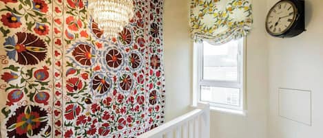 Una habitación con un papel tapiz estampado, un candelabro, un ventanal con un adornado de flores y un reloj de pared.
