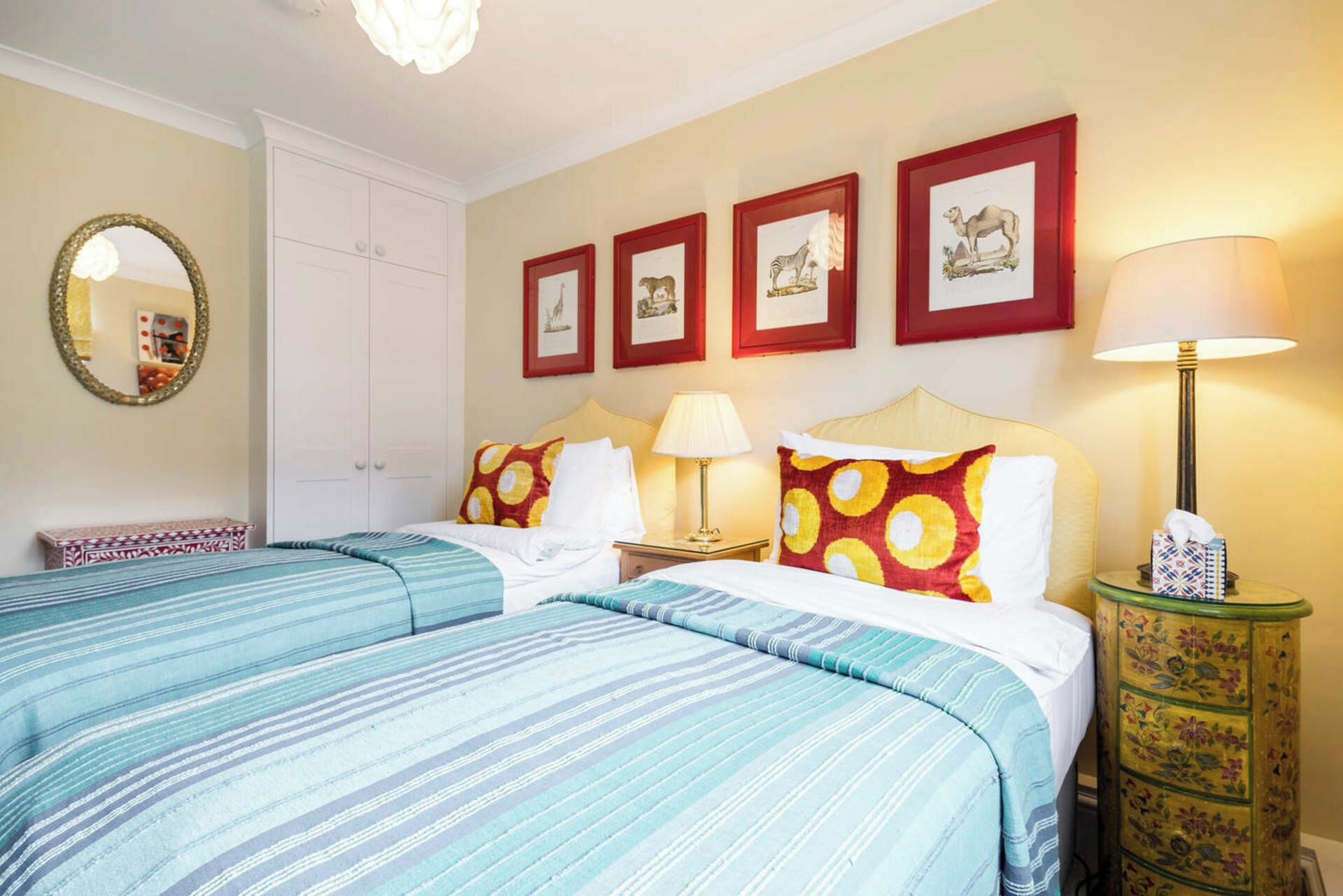 Habitación de hotel con dos camas, una mesita de noche con lámpara, un espejo y cuadros en la pared.
