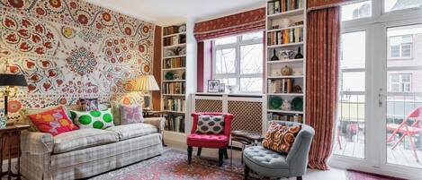 A cozy living room with a sofa, armchairs, a patterned rug, a bookshelf, and a large window with curtains.