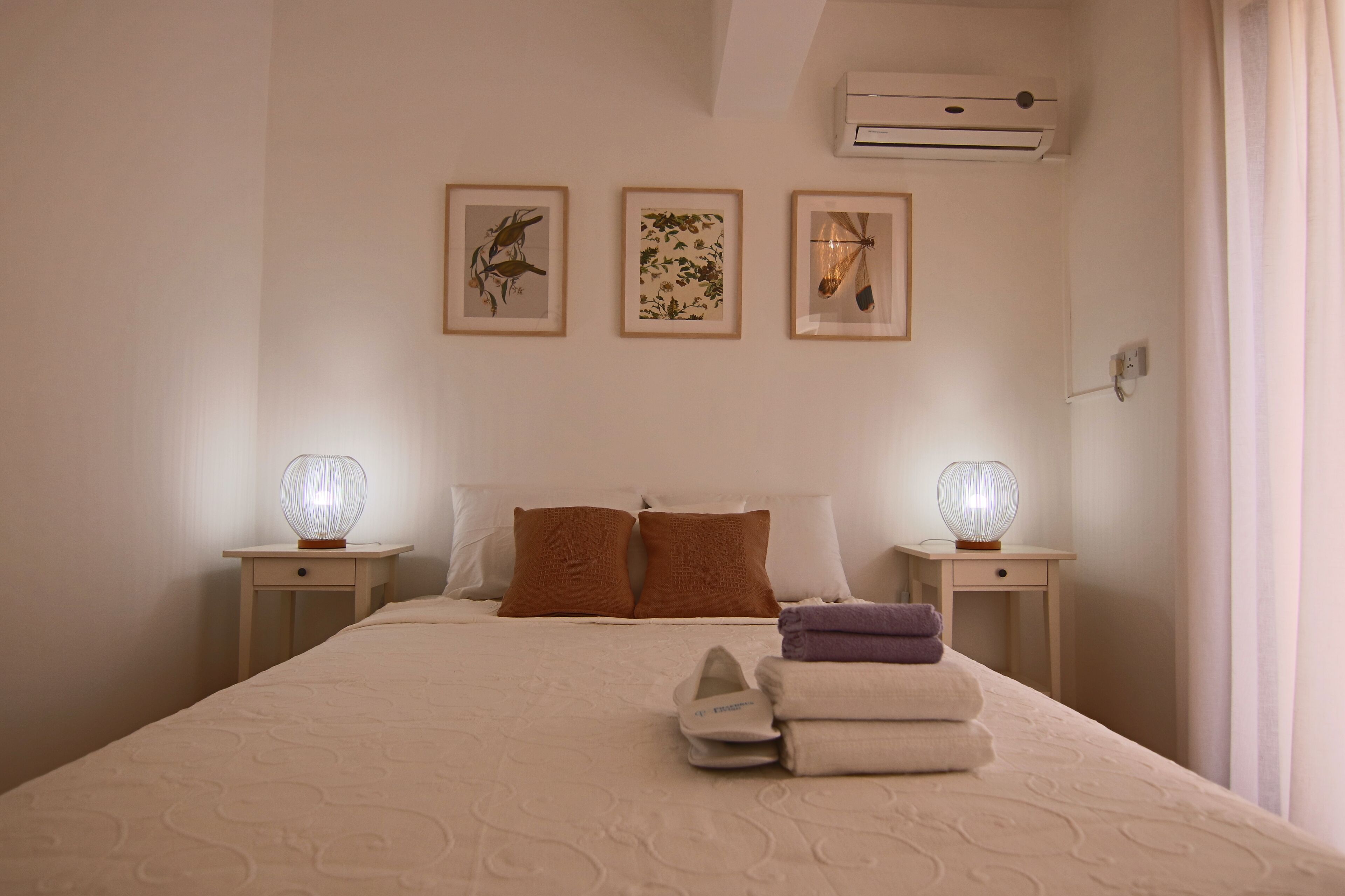 A hotel room with a bed, two bedside tables with lamps, a wall-mounted air conditioner, and three framed pictures.
