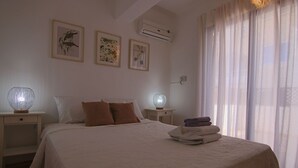 A neatly made bed with a white bedspread, two brown pillows, and a folded towel on the side. There are two bedside tables with lamps, a framed picture on the wall, and an air conditioning unit above the window. - Phaedrus Living: Luxury Flat Protaras Sea View 9 (Paralimni)