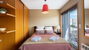 A hotel room with a bed, a red pendant light, a view of the city through a large window, and a wooden wardrobe.