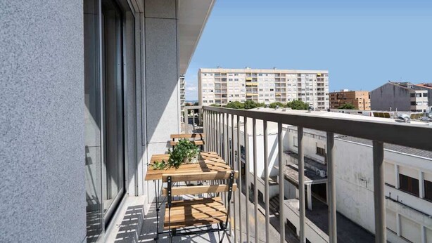 A balcony with a wooden table and chairs, a potted plant, and a view of surrounding buildings.