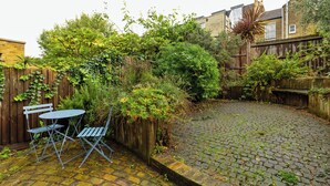 A paved courtyard with a metal table and chairs, surrounded by dense greenery and a wooden fence. - Beautiful 2BD Flat with Private Garden, Clapham (London)