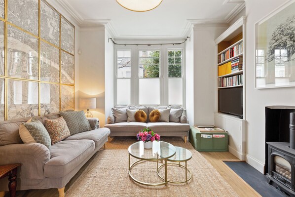 A living room with a sofa, a coffee table, a fireplace, a bookshelf, and a large window with a view of greenery.
