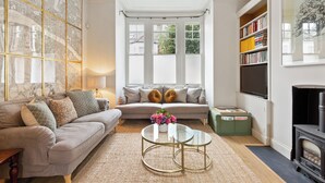 A living room with a sofa, a coffee table, a fireplace, a bookshelf, and a large window with a view of greenery. - Beautiful 2BD Flat with Private Garden, Clapham (London)