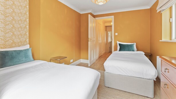 A hotel room with two beds, a small table, a dresser, and a hallway leading to another room. - Beautiful 2BD Flat with Private Garden, Clapham (London)