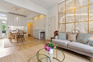 A modern living room with a sofa, a glass coffee table with flowers, and a dining area with a table and chairs. - Beautiful 2BD Flat with Private Garden, Clapham (London)