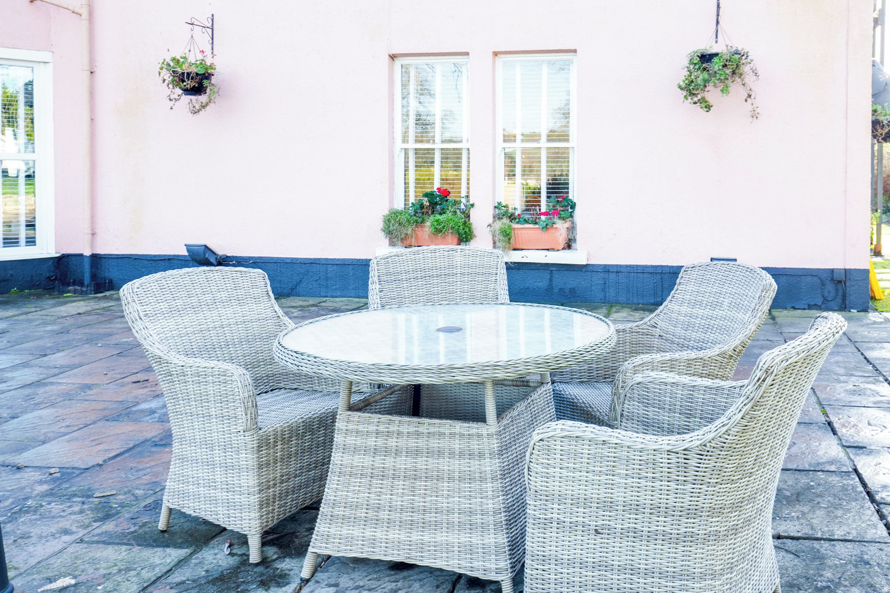 A round wicker table with six chairs, potted plants on the wall, and a window with blinds.