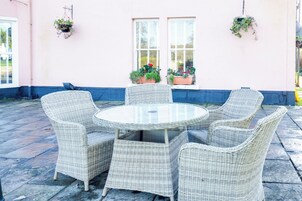 Una mesa redonda de mimbre con seis sillas, plantas en macetas en la pared y una ventana con persianas.