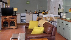 Ein gemütliches Wohnzimmer mit einer braunen Ledersofa, einem Holztisch und einem Fernseher auf einem Holzkasten. Im Hintergrund ist eine Küche mit weißen Schränken und einem Spülbecken zu sehen.