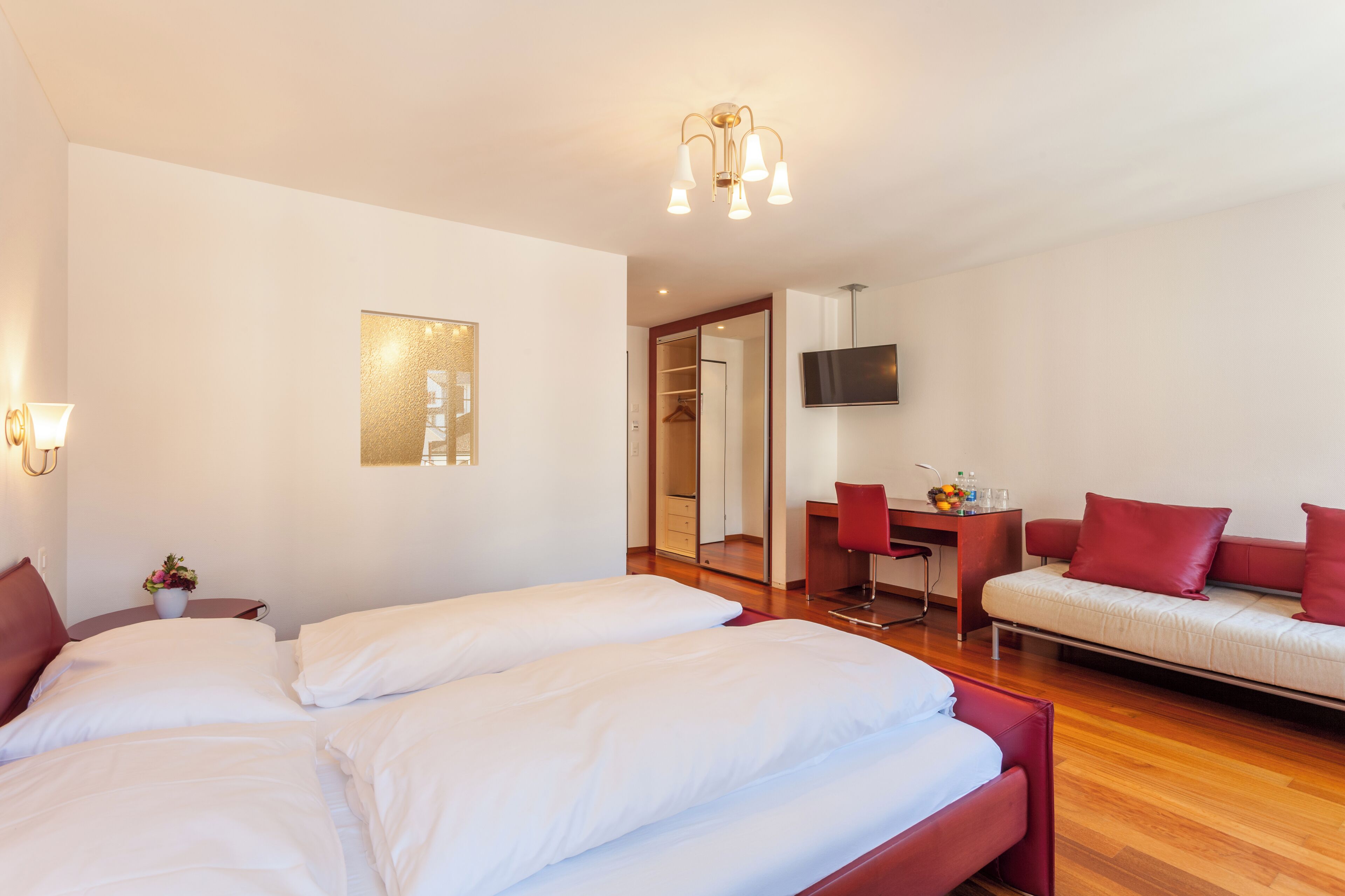 A hotel room with a large bed, a sofa, a desk with a chair, a TV, and a mirror.
