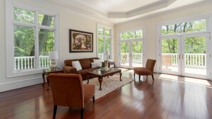 A spacious living room with hardwood flooring, a brown sofa set, a coffee table, and large windows offering a view of the outdoors. - Wedding Venue - Maison D'artagnan on Pimmit Run - Great for Extended Stays (McLean)