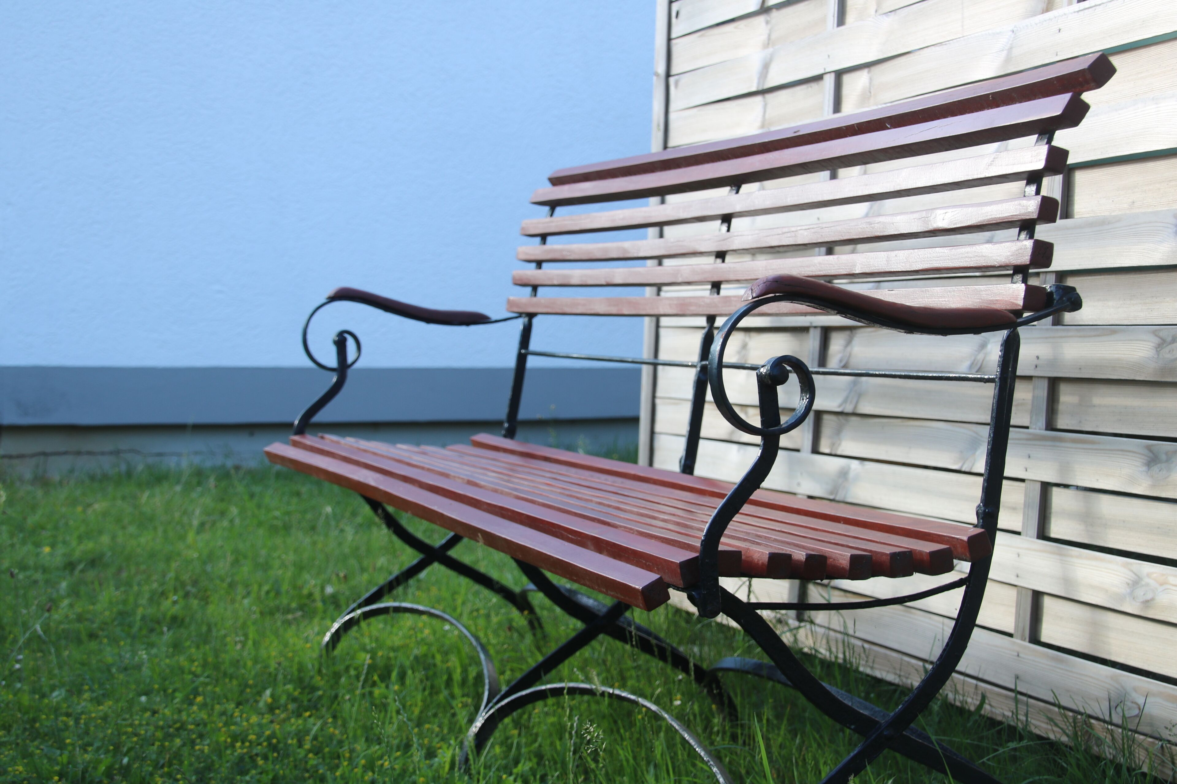 A wooden bench with metal armrests and legs, positioned on grass in front of a wooden building.