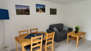 A living room with a wooden dining table, two chairs, a grey sofa, and a small side table with a plant. There are three framed pictures on the wall. - Vacation apartment "Eselstube"! Vacation on a farm in the Eifel (Nettersheim)