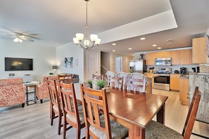 A modern kitchen with built-in appliances, a dining area with a wooden table and chairs, and a living room with a flat-screen TV and patterned sofas.