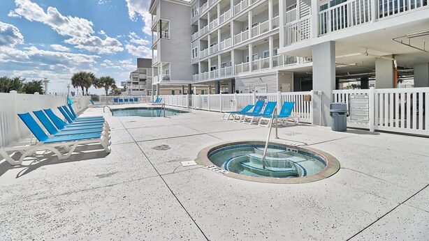 Zona de piscina con sillones de descanso y bañera de hidromasaje, rodeada por una cerca blanca y un edificio de varios pisos con balcones.