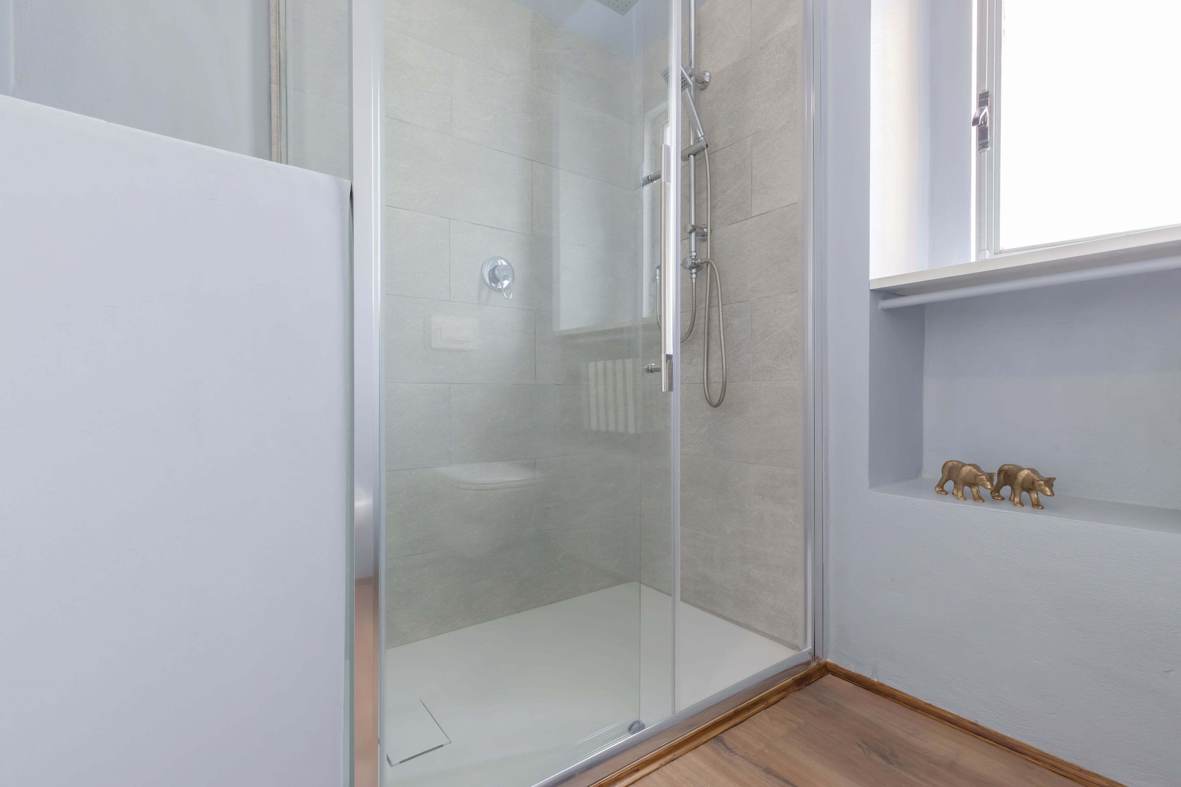 A glass-enclosed shower with a white tiled wall, a wooden floor, and a small shelf with two golden animal figurines.