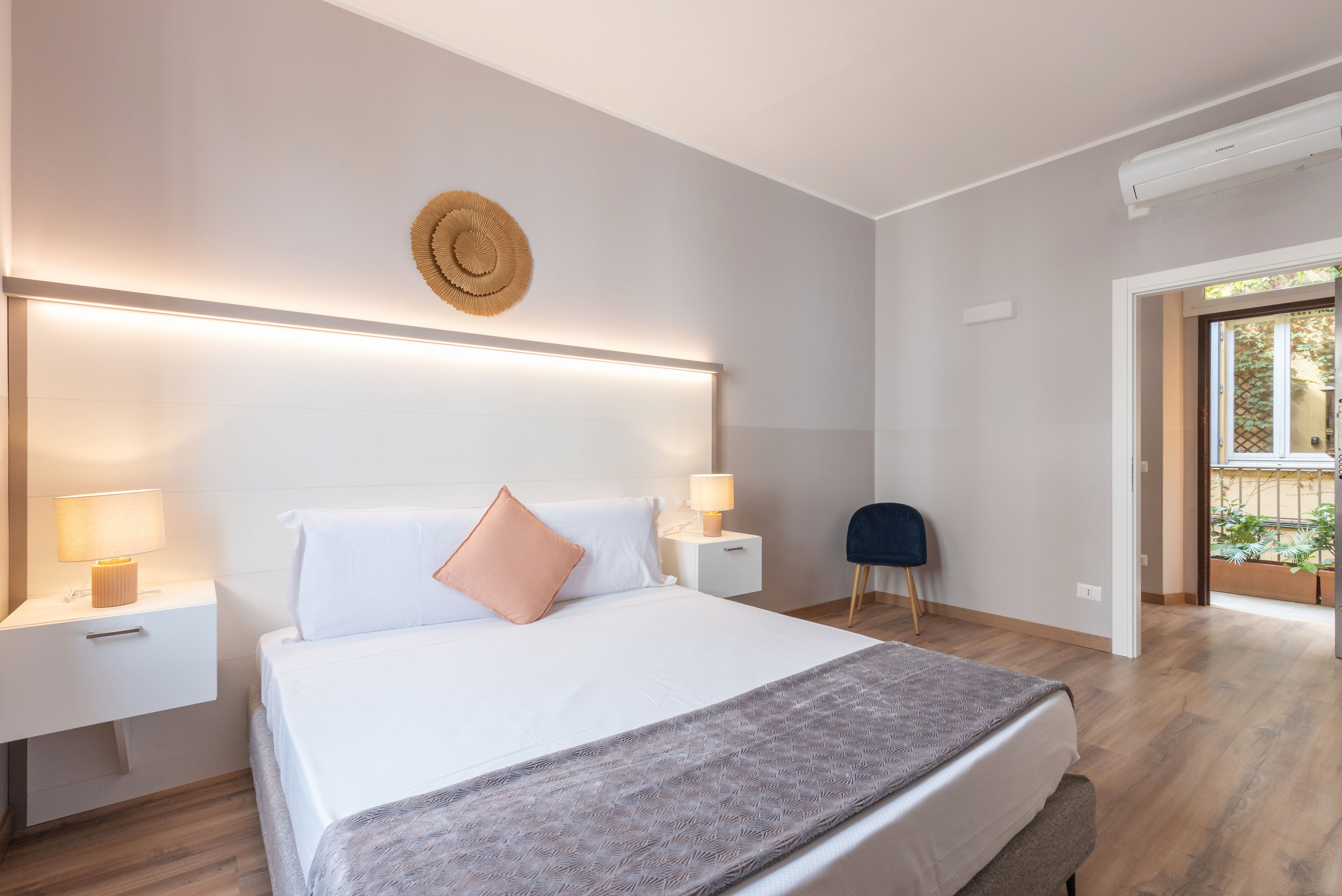 A modern bedroom with a large bed, bedside tables with lamps, a round wall decoration, and a view of the outdoors.