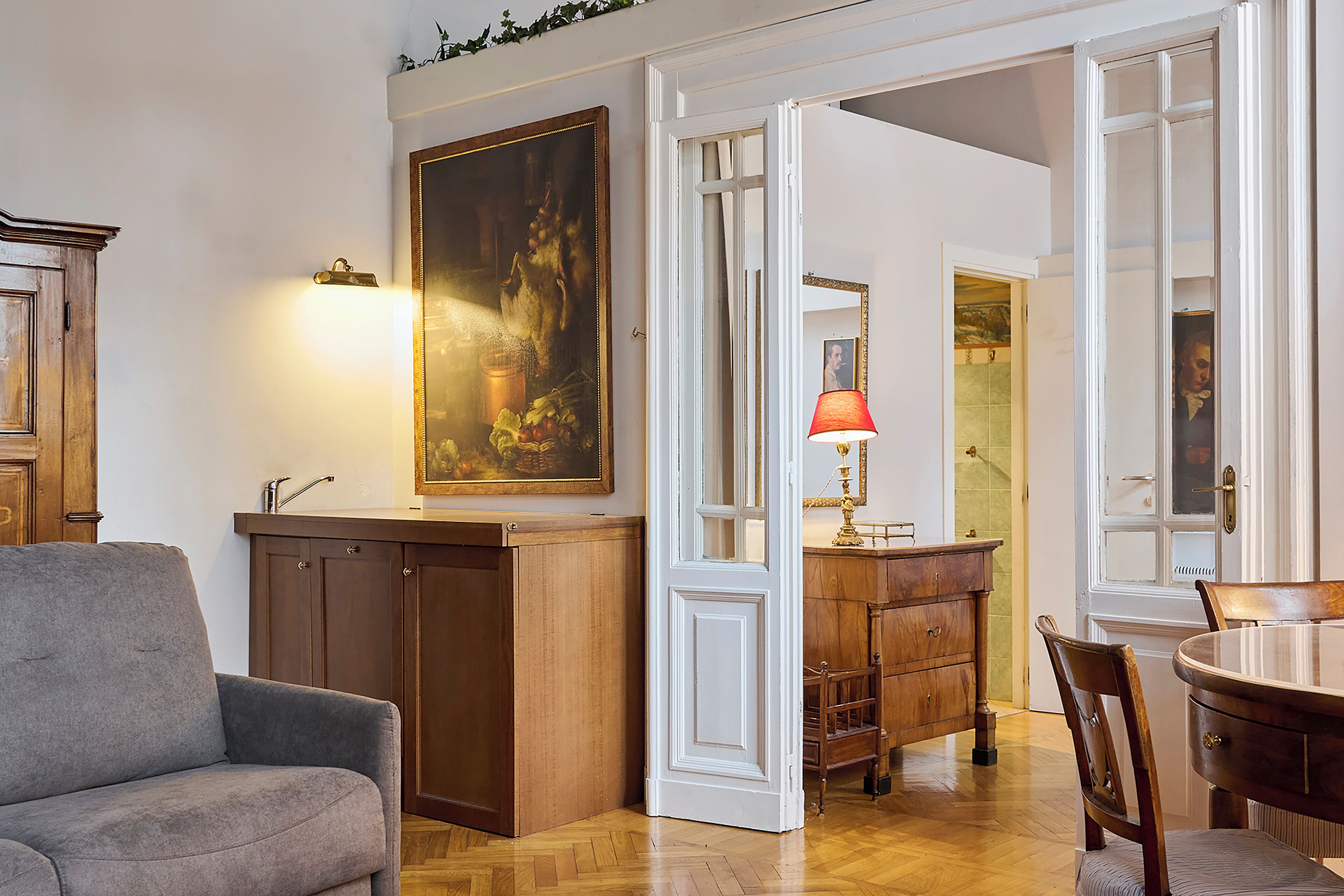 Una habitación con un armario de madera, un sillón gris, una mesa de comedor con sillas y un cuadro en la pared.