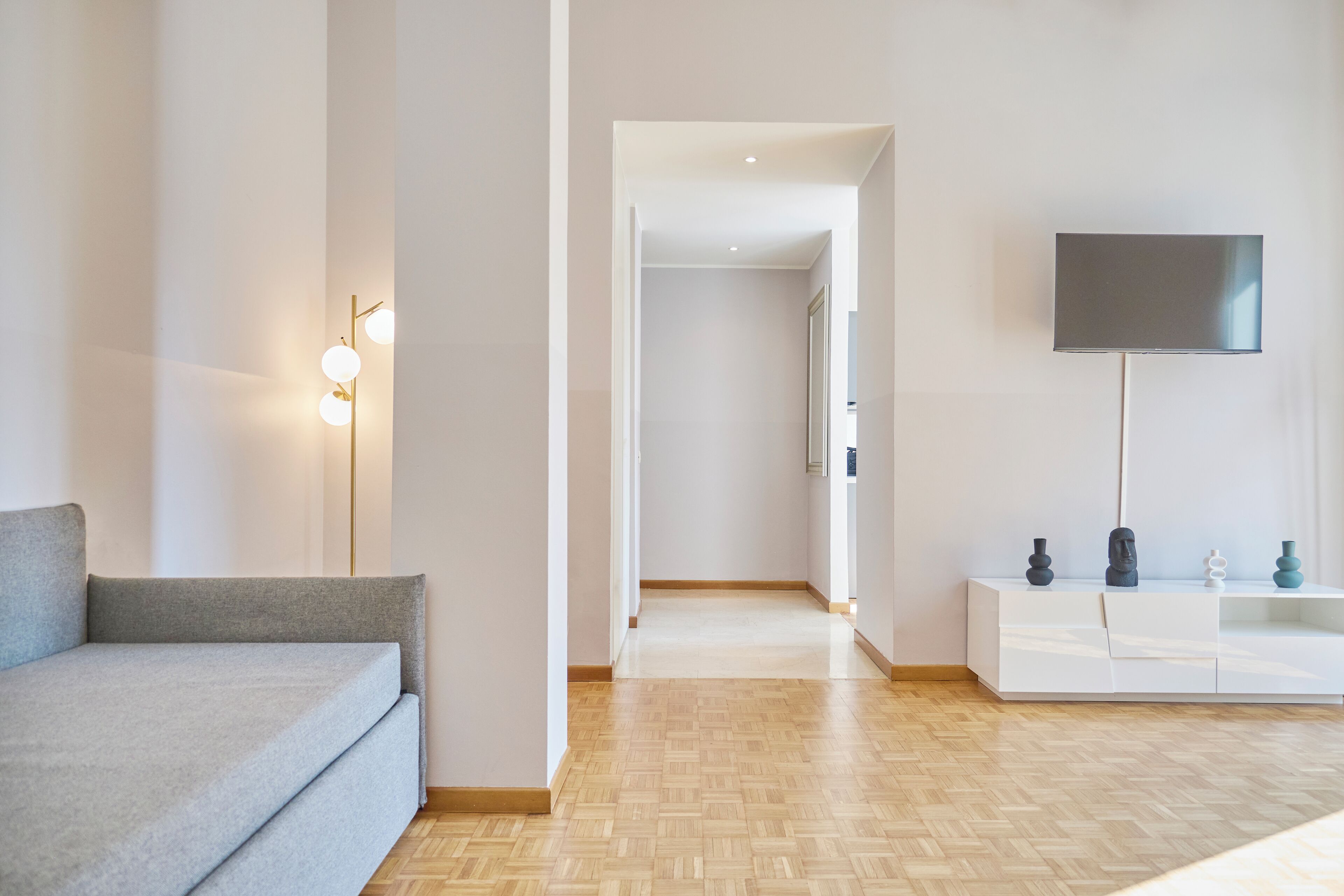 Una sala de estar moderna y minimalista con un sofá gris, piso de madera, una consola blanca con objetos decorativos y un televisor de pantalla plana montado en la pared.