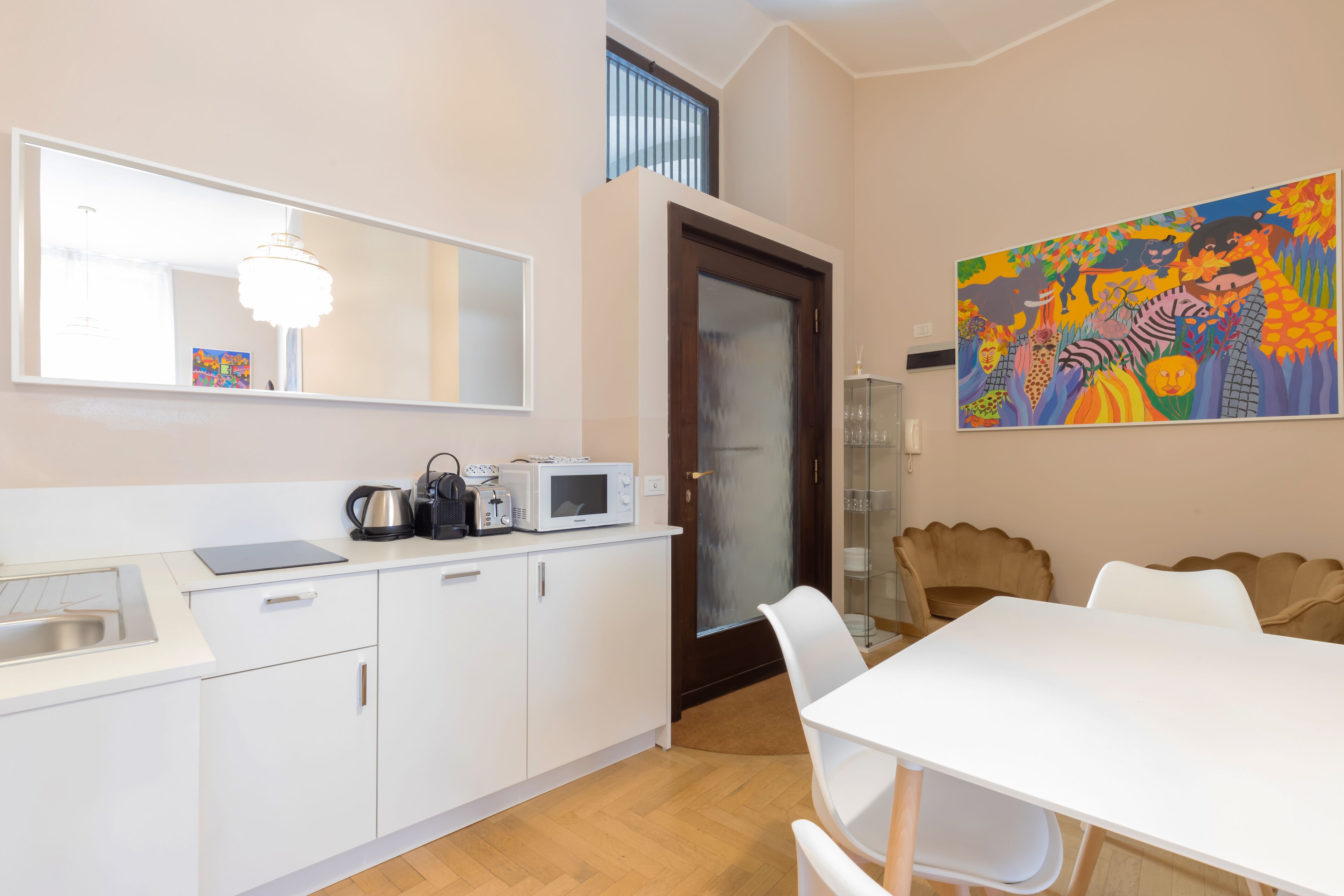 Una cocina moderna con armarios blancos, un microondas y una tetera. Hay un comedor con una mesa y sillas blancas. Un cuadro colorido adorna la pared.