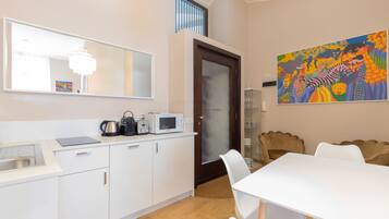 Una cocina moderna con armarios blancos, un microondas y una tetera. Hay un comedor con una mesa y sillas blancas. Un cuadro colorido adorna la pared.