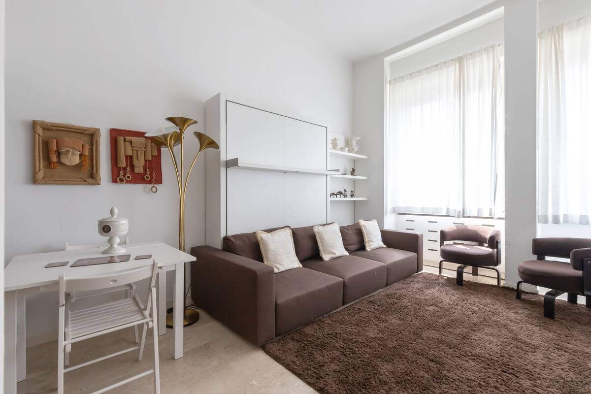 Un salón moderno con un escritorio blanco, un sofá marrón y un ventanal con cortinas translúcidas.