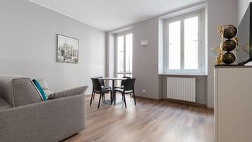 Una sala de estar moderna con un sofá gris, una mesa de comedor con cuatro sillas y un amplio ventanal que proporciona luz natural.