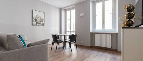 Una sala de estar moderna con un sofá gris, una mesa de comedor con cuatro sillas y un amplio ventanal que proporciona luz natural.