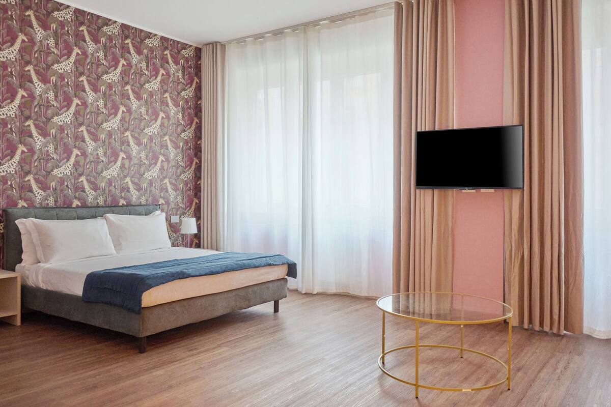 Una habitación de hotel con cama, televisor montado en la pared, una mesa redonda de vidrio y papel tapiz estampado.