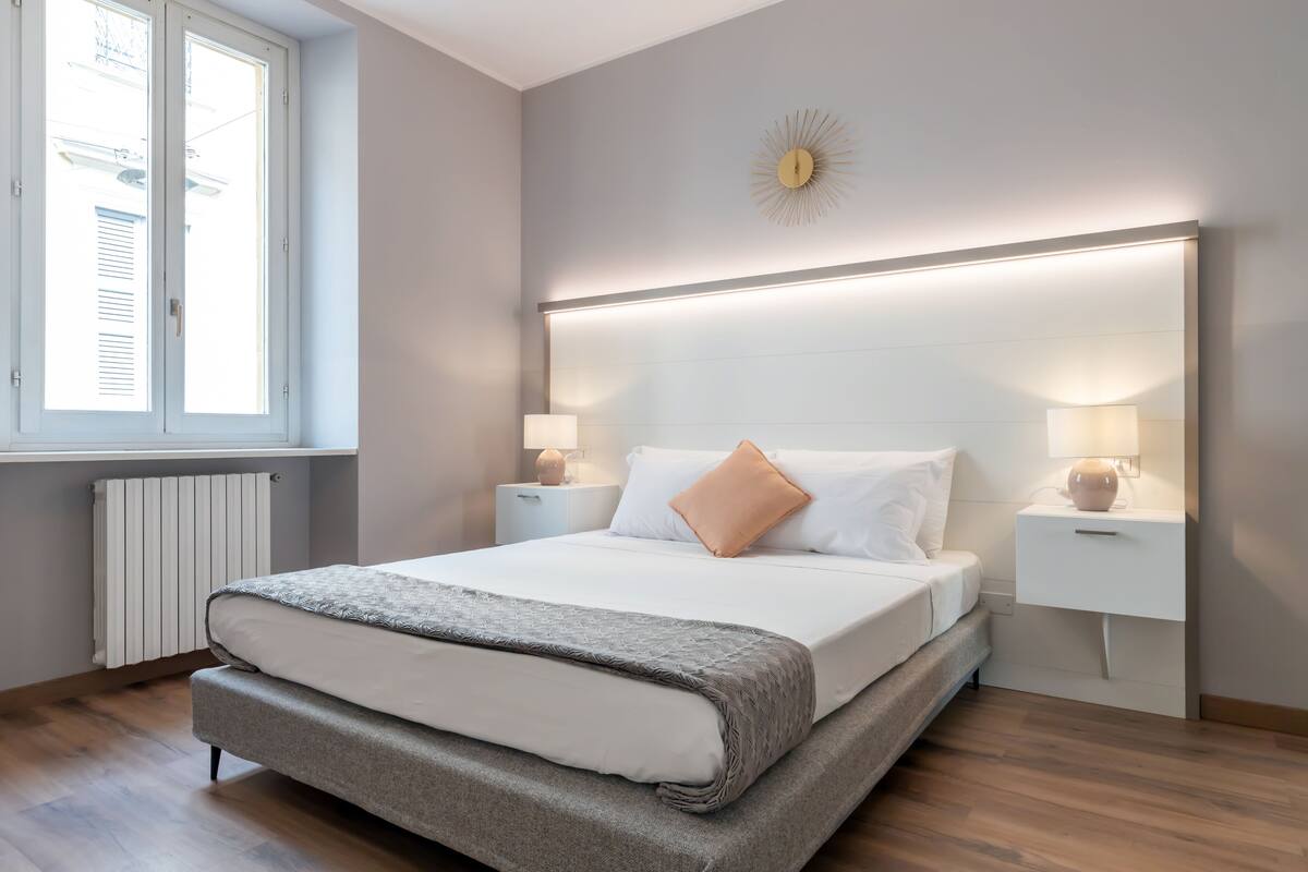 Un dormitorio moderno con una cama grande, lámparas de noche, un reloj de pared y una ventana con persianas.