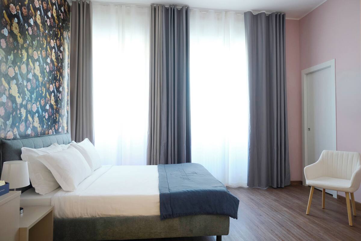 Habitación de hotel con cama, una silla, una mesita de noche y un ventanal con cortinas.