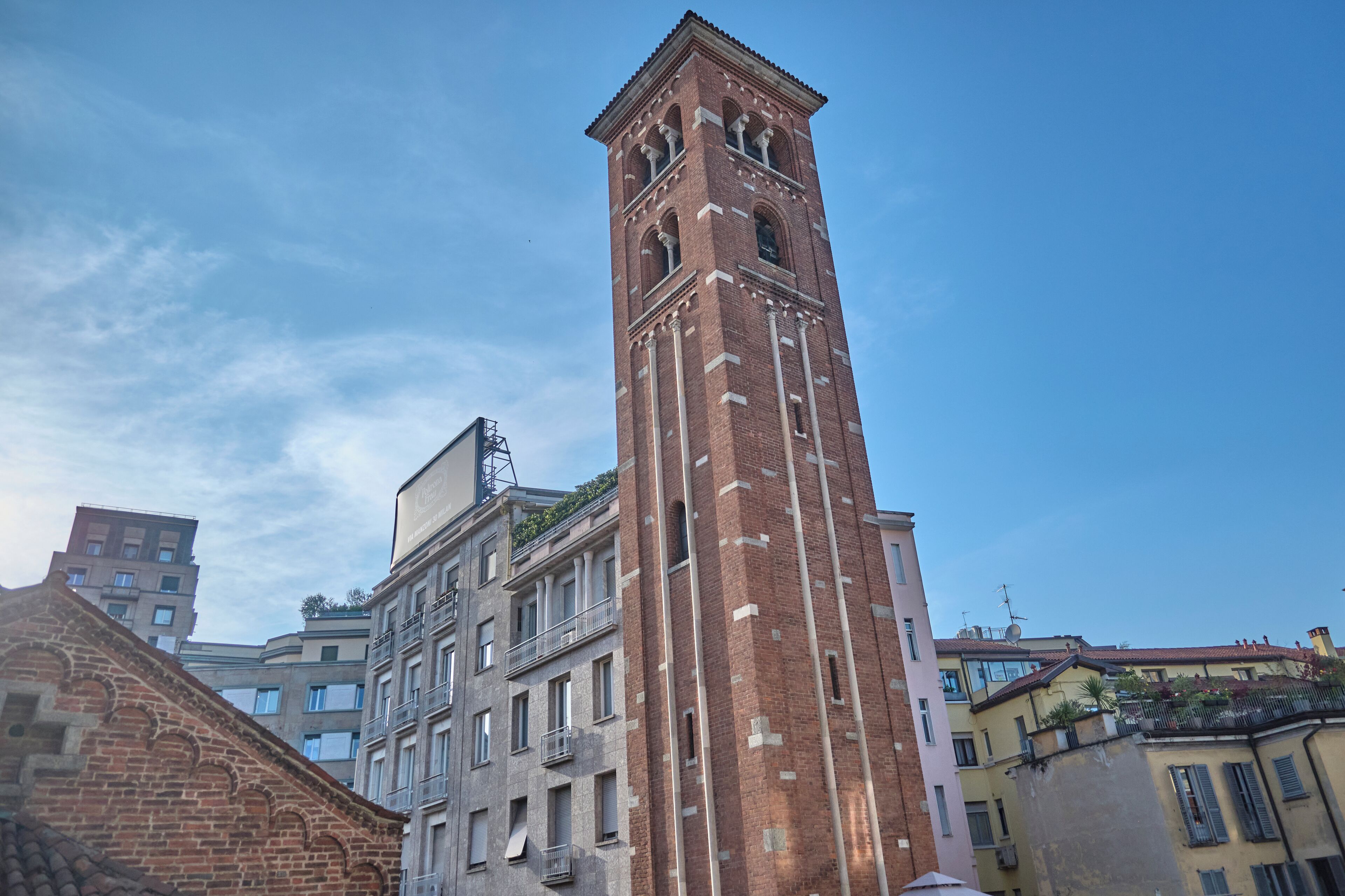 A tall brick tower with arched windows and a balcony, surrounded by multi-story buildings with balconies and windows.