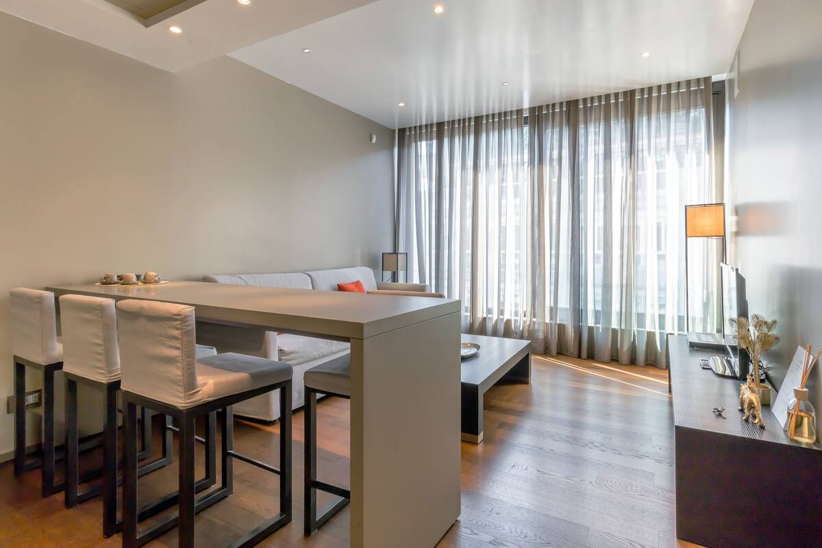 Una habitación de hotel moderna con comedor, sofá, mesa de centro y un amplio ventanal con cortinas.