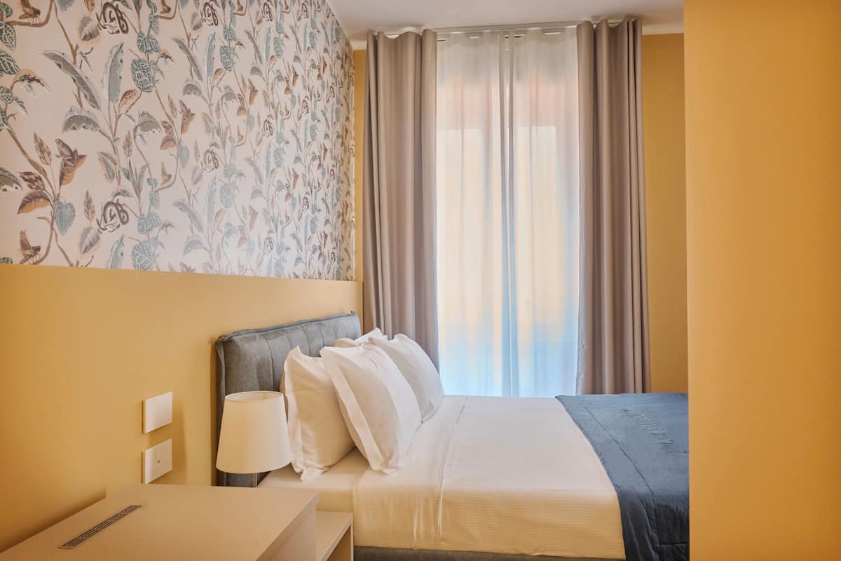 Una habitación de hotel con una cama, una mesita de noche, una lámpara y una ventana con cortinas.